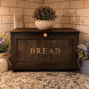 May include: A dark brown wooden bread box with the word "BREAD" in gold lettering. The box has a hinged lid and a small gold knob. Decorative plants in pots and a watering can are on the countertop.