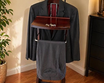 May include: A dark wood valet stand with a round mirror, a jacket hanger, a small shelf, and a trouser bar. A dark gray suit jacket and trousers are displayed. The shelf holds cufflinks. The text on the wall says "Stylish Functional Elegant."