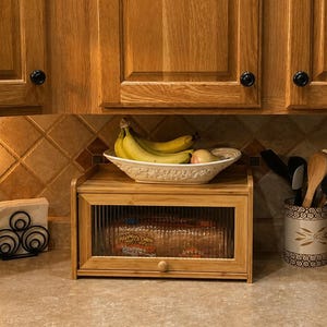May include: A wooden bread box with a glass front sits on a countertop, next to a decorative frame and a napkin holder. A bowl of bananas and fruit sits above the bread box. Kitchen utensils are in a decorative holder.