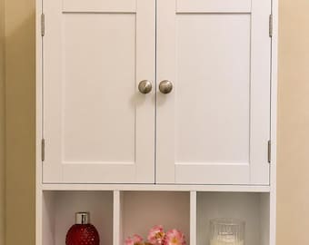 May include: White wall-mounted cabinet with two doors and silver knobs. Below, there are three open cubbies holding a red decorative bottle, pink flowers, and a candle. A small plant and diffuser sit on top. Two rolls of toilet paper are in a basket below.