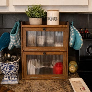 May include: A two-tiered wooden cabinet with glass doors, holding dishes. A ceramic jar with "GOOD DOG" text sits on top, along with a small plant. Blue and white checkered oven mitts hang on either side. A decorative pig and tray are on the counter.