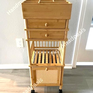 May include: A light brown bamboo storage cart with multiple compartments and drawers. The top section has several open slots, followed by two drawers with round knobs. The bottom features a woven rattan panel and sits on wheels.