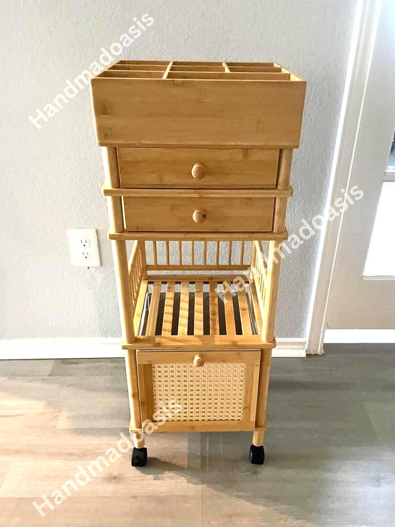 May include: A light brown bamboo storage cart with multiple compartments and drawers. The top section has several open slots, followed by two drawers with round knobs. The bottom features a woven rattan panel and sits on wheels.
