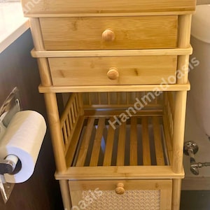 May include: A light brown bamboo bathroom storage unit with a top shelf, two drawers, and a lower shelf with a woven panel door. The unit has a natural wood finish and round wooden knobs.