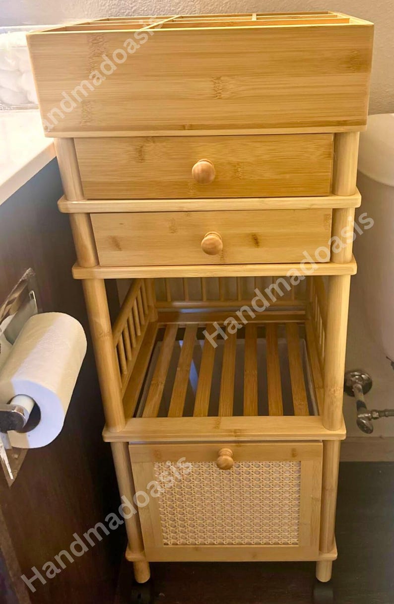 May include: A light brown bamboo bathroom storage unit with a top shelf, two drawers, and a lower shelf with a woven panel door. The unit has a natural wood finish and round wooden knobs.