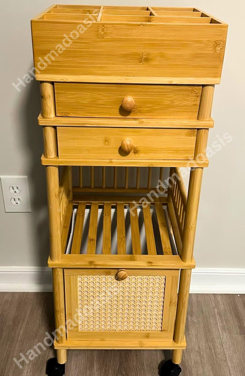 May include: A bamboo storage cart with multiple compartments and drawers. The top section has open dividers, while the middle features two drawers with wooden knobs. The bottom section has a door with a woven panel. The cart is on wheels.