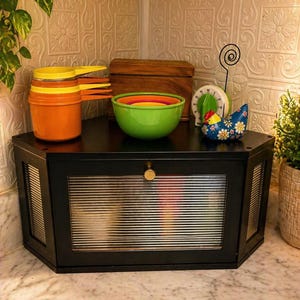 May include: A black, hexagonal bread box with a glass front and gold knob sits on a marble countertop. The box is surrounded by kitchen items, including orange and yellow measuring cups, a wooden box, and a green bowl.