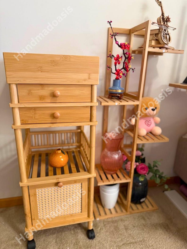 May include: A light brown bamboo storage cabinet with two drawers, a slatted shelf, and a woven door. A matching bamboo plant stand with multiple shelves displays decorative items, including a vase with red flowers, a teddy bear, and a decorative violin.