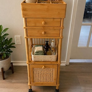 May include: A light brown bamboo rolling cart with multiple storage compartments. The top section has dividers for organizing items, with two drawers below. A woven basket and a cabinet with a woven front are also included.