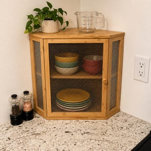 May include: A bamboo corner cabinet with wire mesh doors, holding colorful bowls and plates. A clear measuring cup and a potted plant sit on top. Salt and pepper grinders are on the countertop.