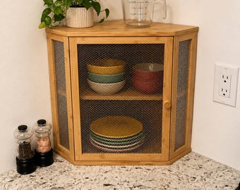 May include: A bamboo corner cabinet with wire mesh doors, holding colorful bowls and plates. A clear measuring cup and a potted plant sit on top. Salt and pepper grinders are on the countertop.
