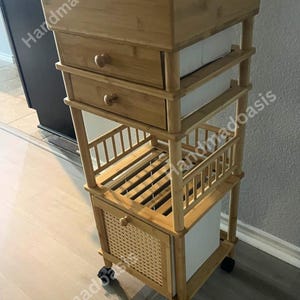 May include: A bamboo rolling cart with multiple storage compartments. The cart features a top section with dividers, two drawers with wooden knobs, and a lower cabinet with a woven front. The cart is on wheels for easy mobility.