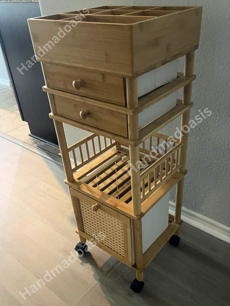May include: A bamboo rolling cart with multiple storage compartments. The cart features a top section with dividers, two drawers with wooden knobs, and a lower cabinet with a woven front. The cart is on wheels for easy mobility.