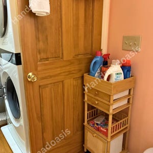 May include: A wooden laundry cart with multiple shelves and a drawer, holding laundry detergents and dryer sheets. The cart is on wheels and is positioned next to a washing machine and a wooden door.