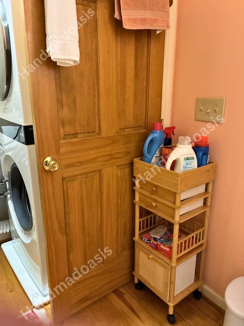 May include: A wooden laundry cart with multiple shelves and a drawer, holding laundry detergents and dryer sheets. The cart is on wheels and is positioned next to a washing machine and a wooden door.
