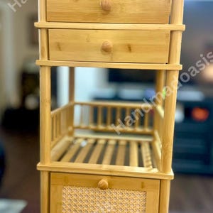 May include: A light brown bamboo storage unit with two drawers, a shelf, and a lower cabinet with a woven rattan panel. The unit has a natural finish and is designed for storage. The drawers and cabinet have round wooden knobs.