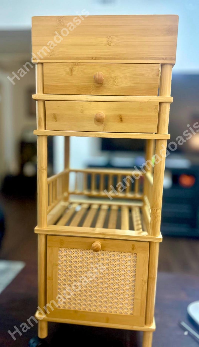 May include: A light brown bamboo storage unit with two drawers, a shelf, and a lower cabinet with a woven rattan panel. The unit has a natural finish and is designed for storage. The drawers and cabinet have round wooden knobs.