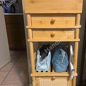 May include: A light-colored bamboo storage cabinet with three drawers and two open shelves. The cabinet has a woven panel on the bottom and wooden knobs. Two blue bags and a white object are on the shelves. The text "Handmadoasis" is visible.