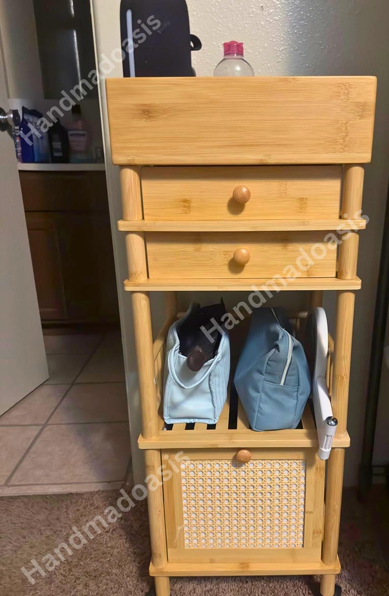 May include: A light-colored bamboo storage cabinet with three drawers and two open shelves. The cabinet has a woven panel on the bottom and wooden knobs. Two blue bags and a white object are on the shelves. The text "Handmadoasis" is visible.