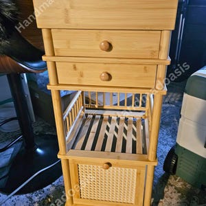 May include: A light-colored bamboo storage cart with two drawers, a slatted shelf, and a woven basket-style drawer. The cart has a rectangular top and is supported by four legs with small wheels.