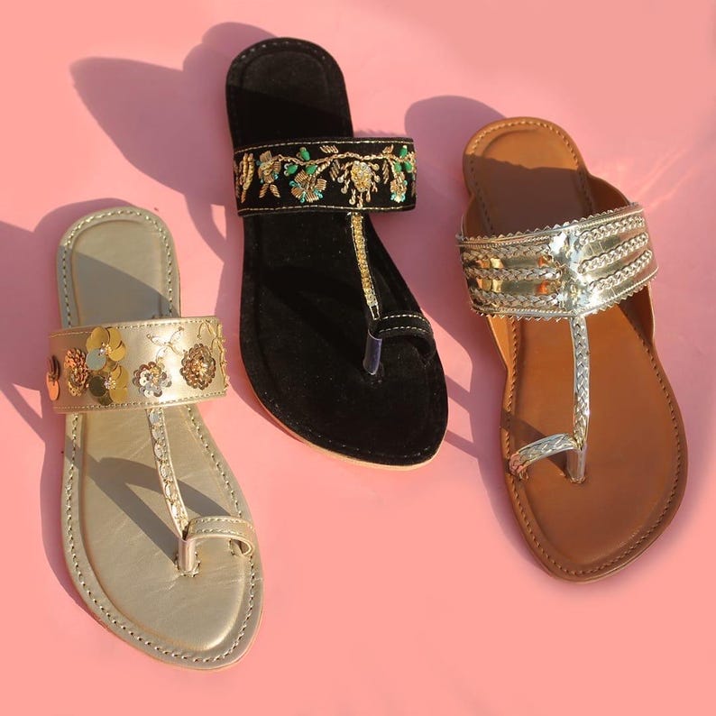 May include: Three pairs of flat sandals in gold, black, and brown, each with unique embellishments. The gold sandal features floral accents, the black sandal has embroidered details, and the brown sandal has silver straps. All have a toe loop design.