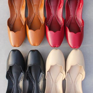 May include: Overhead shot of eight pairs of leather flats in various colors. The colors include tan, red, black, and cream. The shoes have a scalloped edge and are stitched along the seams.
