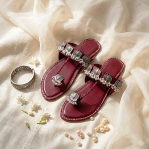May include: A pair of deep red flat sandals featuring a toe ring design, adorned with silver-toned lotus-shaped metal embellishments and small white beads along the straps. A matching silver-toned filigree bangle is positioned to the left.