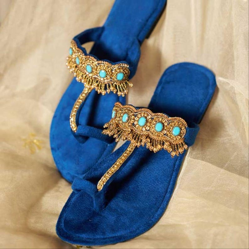 May include: A pair of blue velvet sandals with gold and turquoise embellishments. The sandals feature a thong style design with a decorative band across the top of the foot. The gold embellishments are intricately detailed.