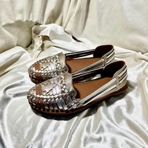 May include: A pair of gold and silver woven leather flats. The shoes have a brown leather insole and a woven design on the toe. The sides of the shoes have white and silver stripes. The shoes are displayed on a white satin surface.