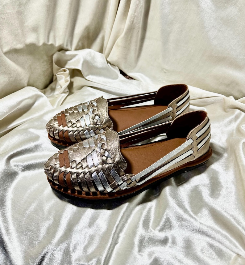 May include: A pair of gold and silver woven leather flats. The shoes have a brown leather insole and a woven design on the toe. The sides of the shoes have white and silver stripes. The shoes are displayed on a white satin surface.