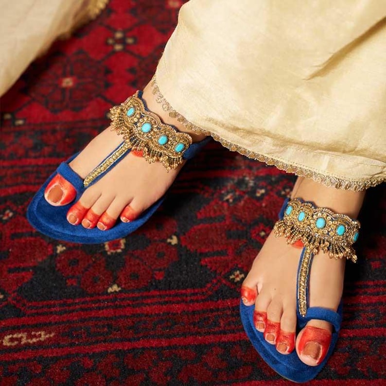 May include: Close-up of blue suede sandals with gold and turquoise anklets. The sandals have a toe loop design. The feet are adorned with red nail polish, and the background is a red patterned rug.