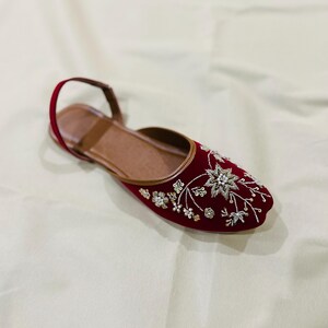 May include: A red velvet flat shoe with intricate floral embroidery in white and gold. The shoe features a brown leather insole and a thin strap around the ankle. The shoe is designed for comfort and style.