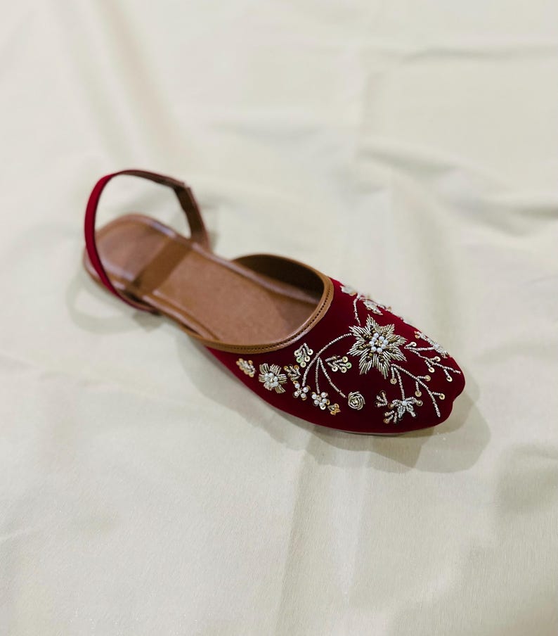 May include: A red velvet flat shoe with intricate floral embroidery in white and gold. The shoe features a brown leather insole and a thin strap around the ankle. The shoe is designed for comfort and style.
