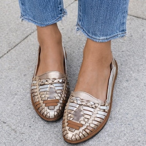 May include: A pair of gold-colored woven leather flats. The shoes have a metallic sheen and a braided detail across the top. The soles are brown, and the shoes are worn with blue denim jeans.