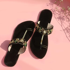 May include: A pair of black sandals with decorative floral embroidery in green, gold, and white. The sandals have a toe loop and a strap across the top of the foot. The background is a solid pink.