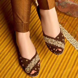 May include: Brown suede peep-toe sandals with gold embroidery. The sandals feature a criss-cross design over the foot and are embellished with floral patterns. The shoes are paired with brown pants, creating a cohesive look.