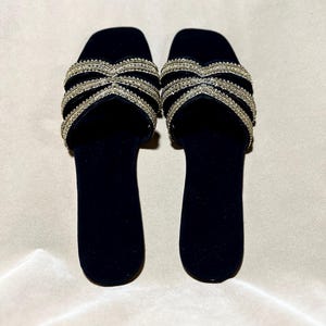May include: A pair of black velvet slide sandals. The sandals feature a crisscross design across the top, embellished with rows of small, sparkling rhinestones. The soles are a solid black, and the sandals are set against a neutral background.