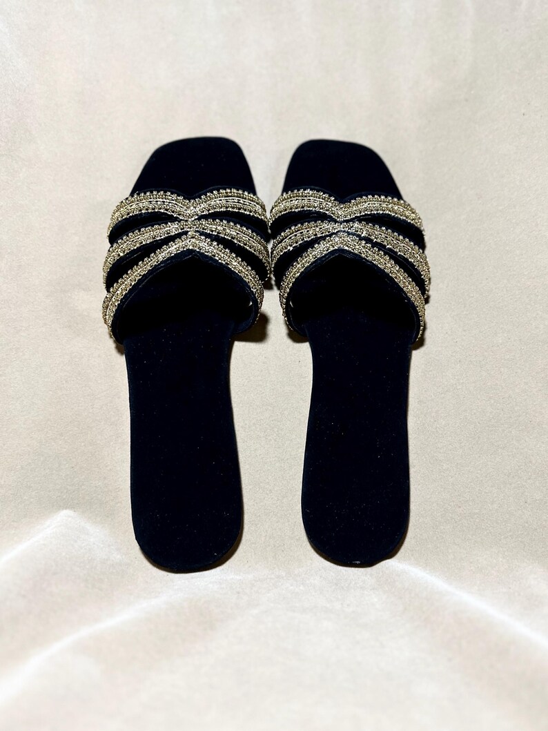 May include: A pair of black velvet slide sandals. The sandals feature a crisscross design across the top, embellished with rows of small, sparkling rhinestones. The soles are a solid black, and the sandals are set against a neutral background.