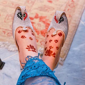 May include: A pair of silver-colored, ornate slip-on shoes with intricate floral and geometric patterns. The shoes have a pointed toe and a decorative design in red and black. The feet are adorned with henna tattoos, and the ankles are wrapped in blue lace.