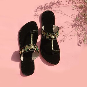 May include: A pair of black sandals with a toe post design. The sandals have a strap decorated with gold, green, and white floral embroidery. The background is a soft pink.