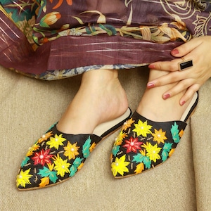 May include: Black slip-on shoes with colorful floral embroidery. The embroidery features yellow, red, green, and orange flowers with white pearl-like accents. The shoes have a pointed toe and a black sole.