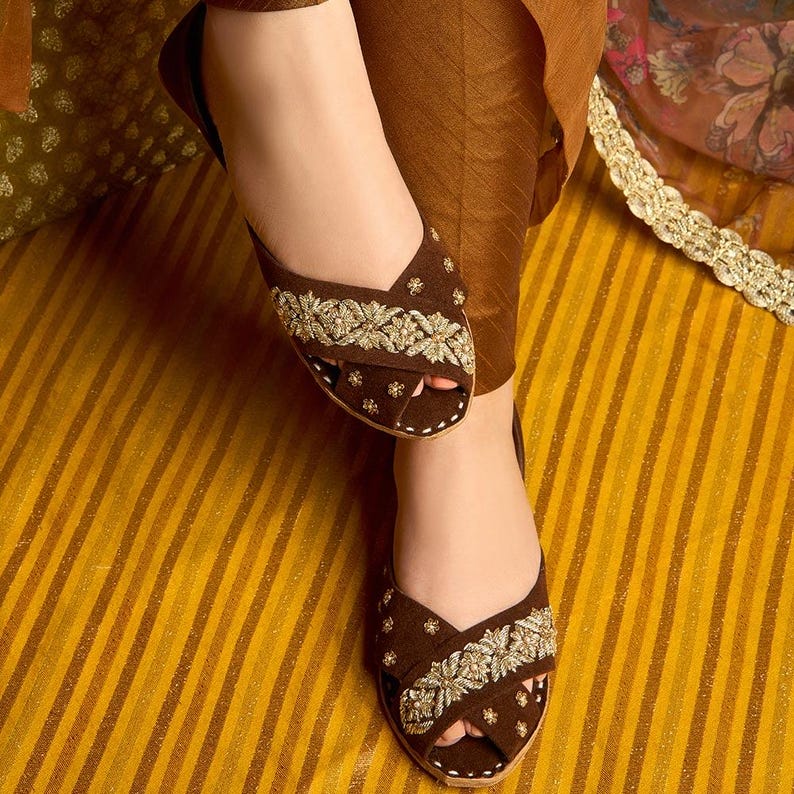 May include: Brown open-toe flats with intricate gold embroidery. The flats feature a criss-cross strap design and are embellished with floral patterns. The shoes are set against a striped yellow and brown background, showcasing a traditional footwear style.