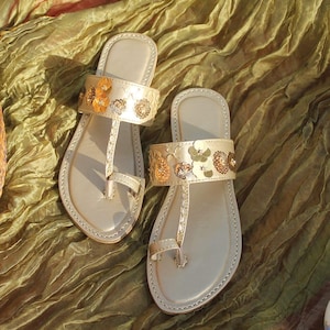 May include: A pair of beige leather sandals with floral embellishments. The sandals feature a thong-style design with a strap adorned with gold and orange flower details. The sandals are displayed on a textured, olive-green fabric.
