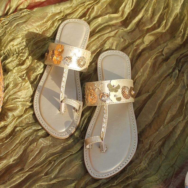 May include: A pair of beige leather sandals with floral embellishments. The sandals feature a thong-style design with a strap adorned with gold and orange flower details. The sandals are displayed on a textured, olive-green fabric.