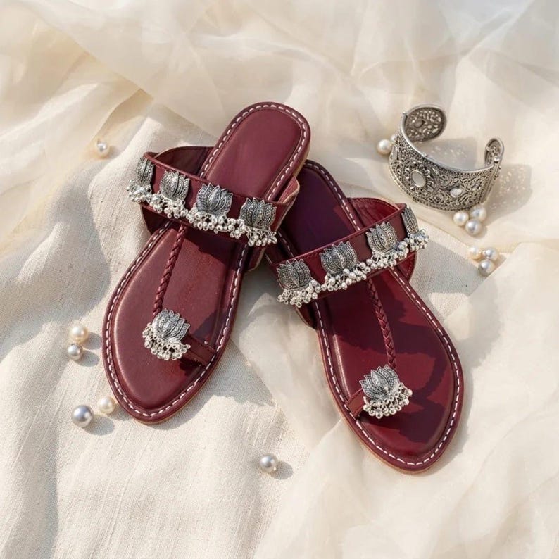 May include: A pair of deep maroon flat sandals featuring a toe ring and a wide strap across the top. Both the toe ring and the strap are adorned with silver-toned metal lotus flower charms and small dangling beads. A matching silver cuff bracelet is included.