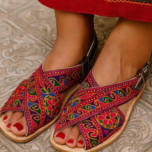 May include: Close-up of colorful embroidered sandals with a cross-strap design. The sandals feature intricate patterns in shades of pink, blue, green, and yellow. The leather soles are light brown, and the straps are dark brown with a buckle.