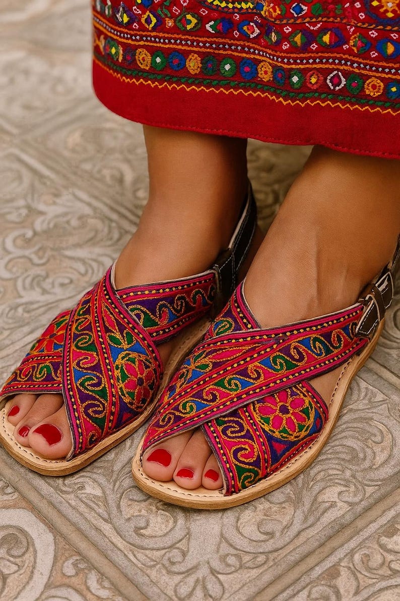 May include: Close-up of colorful embroidered sandals with a cross-strap design. The sandals feature intricate patterns in shades of pink, blue, green, and yellow. The leather soles are light brown, and the straps are dark brown with a buckle.