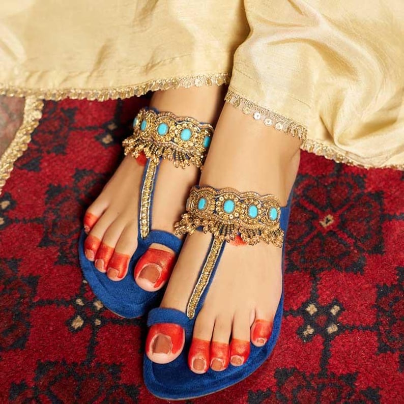 May include: A pair of blue velvet sandals with gold and turquoise anklets. The sandals have a thong design and are adorned with gold embellishments. The toenails are painted red, and the sandals rest on a red patterned rug.