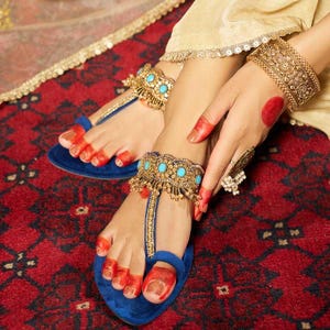 May include: Blue suede sandals with gold and turquoise embellishments. The sandals feature a T-strap design and are adorned with intricate gold detailing. A gold cuff bracelet and anklet with turquoise stones are also visible. The image is set against a red patterned background.