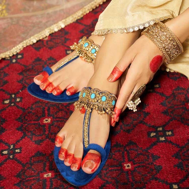 May include: Blue suede sandals with gold and turquoise embellishments. The sandals feature a T-strap design and are adorned with intricate gold detailing. A gold cuff bracelet and anklet with turquoise stones are also visible. The image is set against a red patterned background.